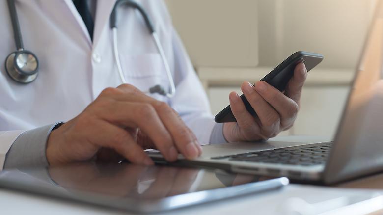 Op de afbeelding is een zorgverlener te zien die een laptop en smartphone gebruikt voor zijn werkzaamheden