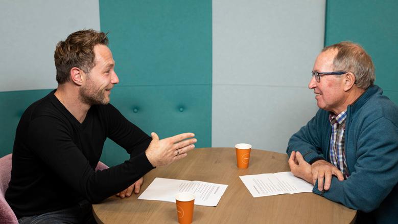 Adviseur patiëntenparticipatie en -communicatie Dennis de Vries en patiënt Hans Barentsen zitten aan een tafel te praten